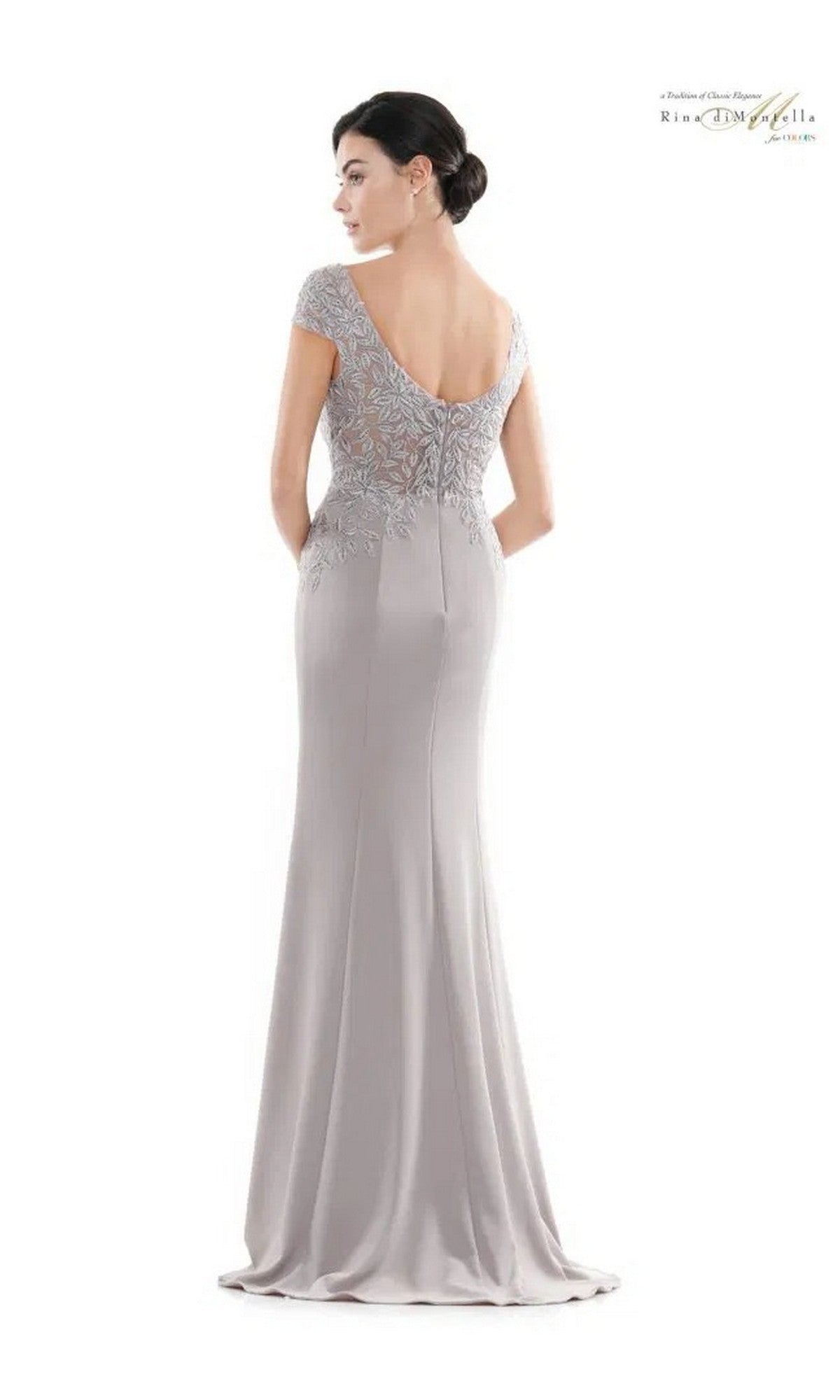A woman models the Rina Di Montella RD2718, a floor-length light gray formal dress with an embroidered bodice, cap sleeves, and a subtle v-neckline, standing with her back to the camera against a plain white background.